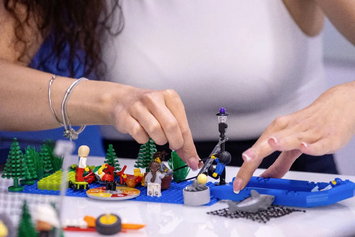 Mâini care ajustează un model construit din piese LEGO pe o plăcuță albastră, într-un workshop LEGO Serious Play.