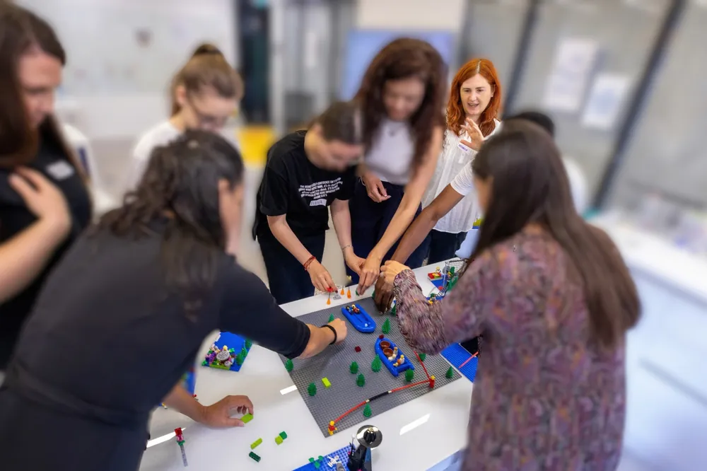 Mihaela Danciu facilitează un grup adunat în jurul unui model comun construit din piese LEGO, pe o masă de lucru.