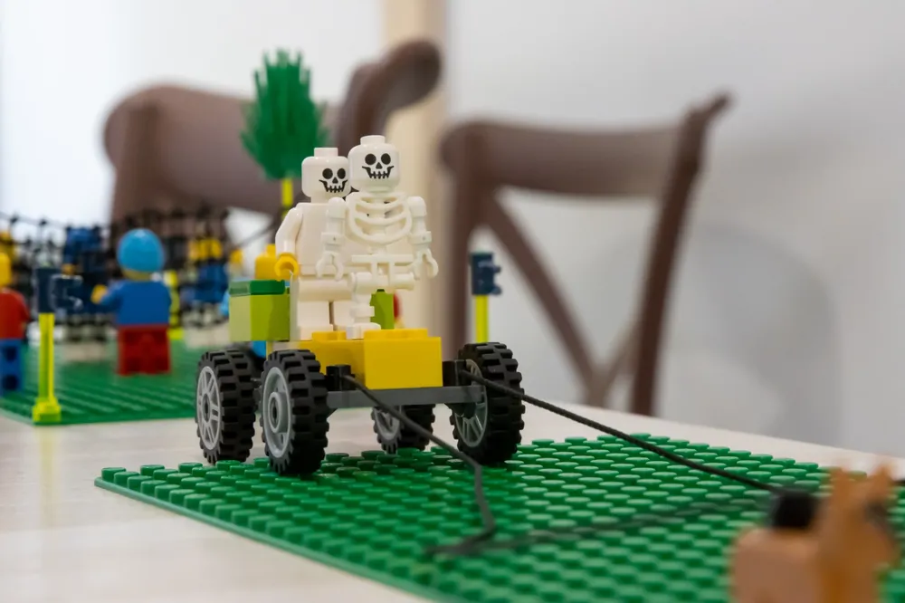 Model din piese LEGO: două figurine scheletice pe o platformă cu roți, trasă cu o sfoară, construit ca metaforă într-un workshop.
